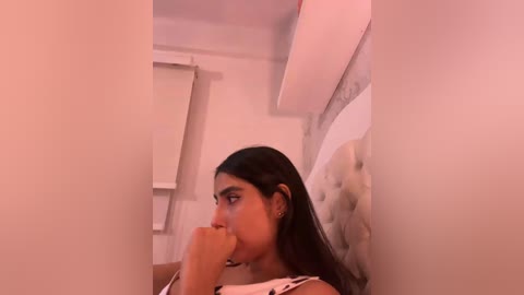 Video of a young South Asian woman with medium brown skin, long black hair, wearing a white sleeveless top, sitting on a tufted white headboard in a cozy, softly lit room with beige walls and a closed white window blind.
