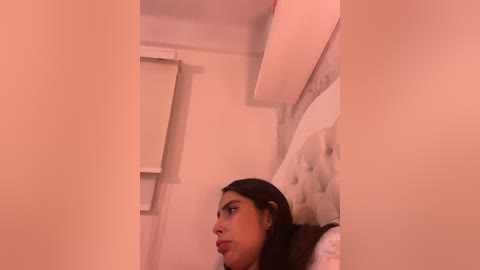 Video of a dark-haired woman with a light skin tone, sitting on a bed with a tufted headboard, in a dimly lit room with soft pink lighting.