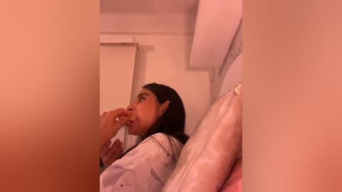 A video of a woman with long dark hair, in a white shirt, eating a sandwich, leaning against a cream-colored wall.