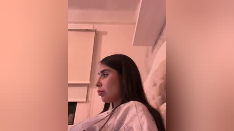 A video of a young woman with long black hair, wearing a white robe, leaning against a beige wall. The background features a closed window with a white blind. The image is dimly lit, creating a soft, muted ambiance.