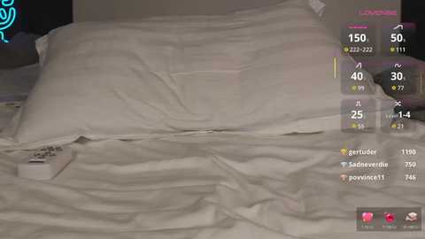 Video of a messy bed with white sheets, a white pillow, and a remote control on the left side. The background shows a virtual screen with a game interface, displaying various statistics and icons.