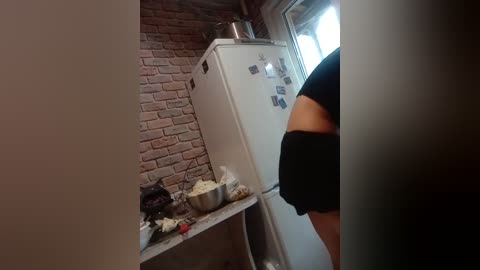 Video of a person in a black shirt standing next to a white refrigerator with photos and a metal pot on top, against a brick wall and countertop cluttered with food items.