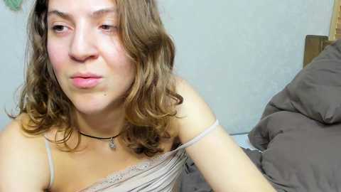 A video of a young woman with light skin and shoulder-length brown hair, wearing a light beige off-shoulder dress, sitting on a bed with brown sheets. She has a neutral expression and a small black pendant necklace.
