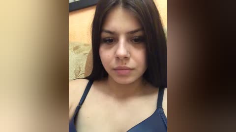 A video of a young Latina woman with straight, dark brown hair and medium skin tone, wearing a dark blue spaghetti-strap top, standing in front of a beige wall with a blurred background.