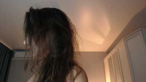 A video of a woman with long, messy black hair in a dimly lit room. She has a pale complexion and is wearing a light-colored top. The background includes a white ceiling fan and a white wardrobe.