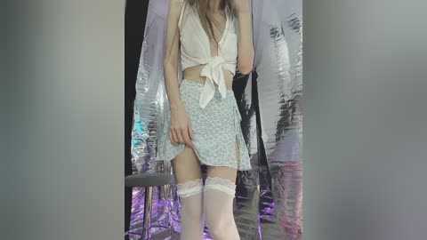 Video of a slender, light-skinned woman with long brown hair, wearing a white crop top, a blue floral skirt, white thigh-high stockings, and a black jacket, standing in front of a silver backdrop.