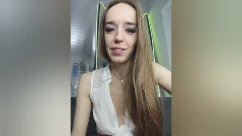 Video of a young Caucasian woman with long, straight light brown hair, wearing a white sleeveless blouse with a low neckline. She's smiling softly, standing indoors with a blurred cityscape background.