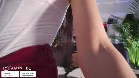 Video of a person in a white and red striped top, bending over, showing a stretch mark on their upper thigh. Background features a green potted plant and a wooden screen.