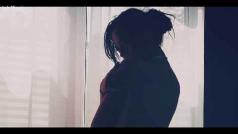 A video depicts a silhouette of a woman with dark hair in a messy bun, standing in front of a window with sheer curtains, casting a shadow.