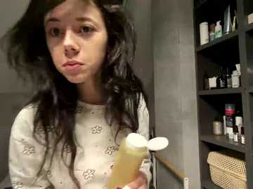 Video of a young woman with long, wavy black hair and fair skin, wearing a white floral-patterned shirt, holding a bottle of clear liquid. Background includes a gray wall and a bookshelf with various toiletries.