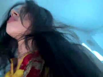 A video of a young woman with long, dark hair blowing wildly, wearing a bright yellow shirt with a red and gold pattern, against a blurry blue sky background.