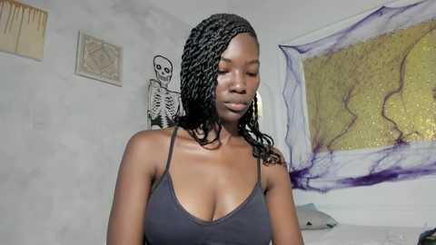 Video of a dark-skinned woman with braided hair, wearing a dark gray spaghetti-strap top, in a room with a wall-mounted skeleton, abstract art, and a white bed.