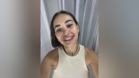 Video of a young woman with short black hair, light skin, and dark eyes, wearing a cream tank top, smiling against a grey curtain backdrop.