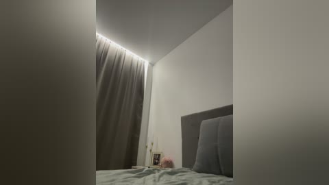 Video of a minimalist, dimly lit bedroom with a bed featuring a grey headboard and a grey and white patterned duvet. The room is dominated by white walls and a sheer curtain draped over the left side, casting a soft glow.