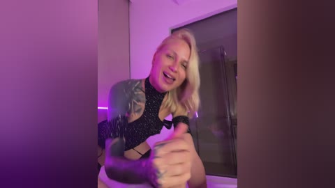 Video of a blonde woman with medium-length hair, wearing a black, glittery top and denim jacket, smiling, holding a drink, in a dimly lit room with purple lighting.
