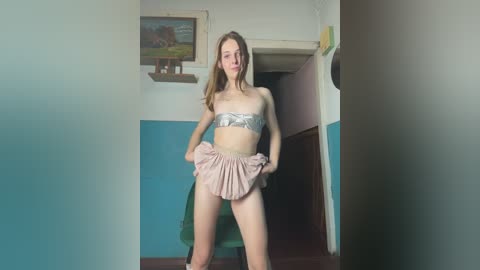 Video of a slender, fair-skinned woman with long, wavy blonde hair, wearing a silver bandeau top and pink pleated skirt, posing seductively in a room with turquoise and white walls, a wooden door, and a framed painting on the wall.