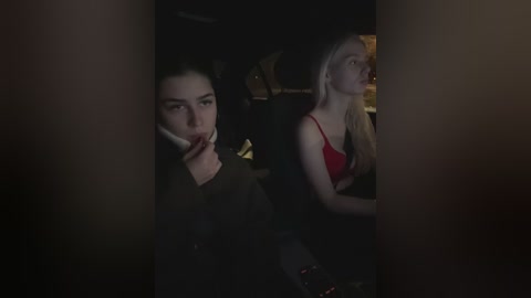 A dimly lit video of two women in a car; one with dark hair, wearing a black coat, and the other with blonde hair, wearing a red top.