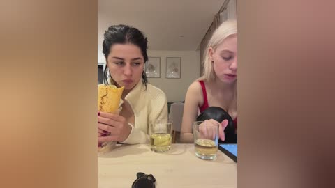 Video: Two women, one with dark hair, one with blonde hair, eating at a table, drinking, in a dimly-lit, modern room.