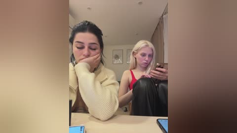 Video of two women in a modern living room. One, with dark hair, rests her head on her hand, wearing a cream sweater. The other, with blonde hair, sits on a black couch in a red tank top, holding a phone.