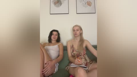 Video of two young women with fair skin, one in a white camisole, the other in a floral dress, sitting on a green couch, surrounded by abstract wall art.