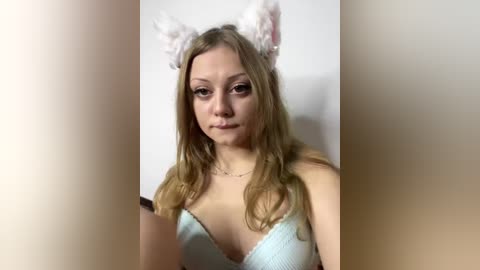 Video of a young, light-skinned woman with long, wavy blonde hair, wearing a light blue bra with white fluffy cat ears. She has a neutral expression, and the background is plain and blurred.