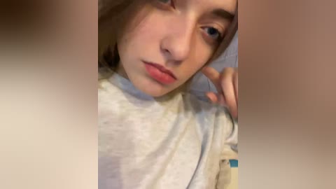 A video of a young Caucasian girl with fair skin, light brown hair, and blue eyes, wearing a gray sweater. She appears relaxed, leaning her head on her right hand, with her left hand slightly visible. The blurred background suggests an indoor setting.