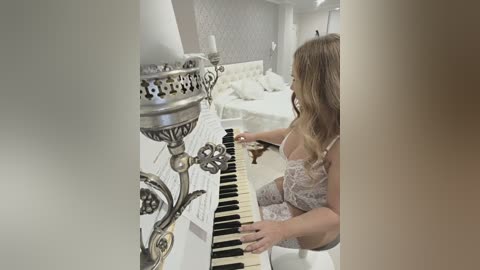 Video of a woman with long, wavy blonde hair, wearing a sheer, lace bra, playing a piano in a luxurious, dimly lit bedroom with ornate silver candleholders on the wall.