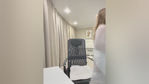 A video of a modern medical examination room with beige walls, light curtains, a gray mesh chair, and a blurred person in a white lab coat.