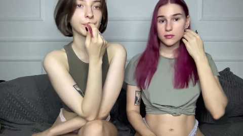 Video of two young women with fair skin, one with short brown hair and the other with long magenta hair, sitting on a gray couch, wearing cropped tops and jeans.