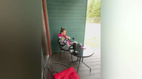 A video of a young child in a pink jacket, sitting on a black wicker chair at a round glass table on a patio, next to a green wooden house.