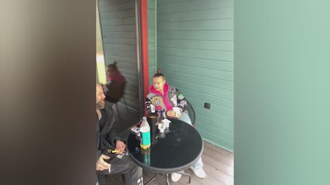 A video of a man in black clothing, a woman in a colorful jacket, and a child in a gray hoodie, sitting at a round glass table in a teal-paneled room with a red pipe.