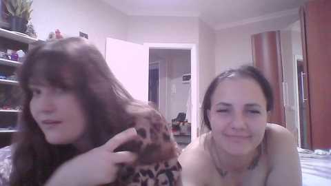 Video of two young women with long hair in a bedroom. One has messy brown hair, the other is brushing it. Background includes shelves, a door, and a mirror.