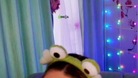 Video of a woman in a playful green frog hat, with pastel blue and green curtains in the background, colorful fairy lights, and a blurred \"DONGA\" logo.