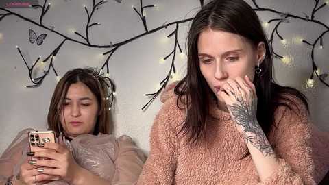 Video of two young women with long hair, one with a pink phone, the other with a tattooed hand on her face, in a cozy room with fairy lights and a tree backdrop.