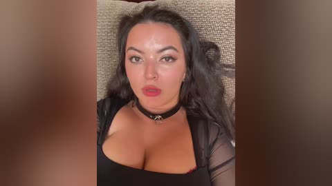 Video of a woman with long black hair, wearing a sheer black top and choker, reclining on a beige couch, lips painted red.