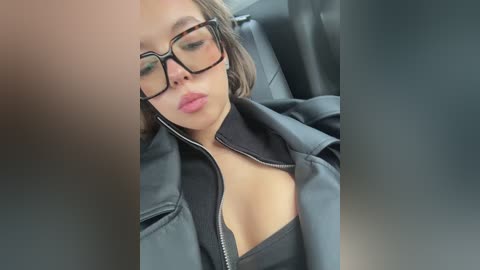 Video of a woman with light brown skin, wearing glasses and a black leather jacket, leaning back in a car seat. She has short, dark hair and her lips are slightly parted.