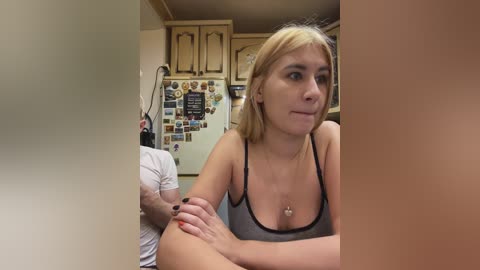 Video of a young woman with fair skin and shoulder-length blonde hair, wearing a gray tank top, sitting in a kitchen with wooden cabinets and a fridge adorned with colorful magnets.