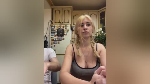 A candid video of a blonde woman in a gray tank top, sitting in a kitchen, with a refrigerator covered in photos and a man in the background.