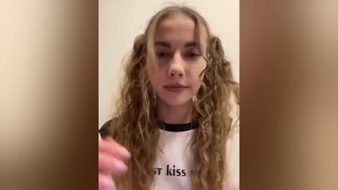 Video of a young woman with long, wavy, blonde hair styled in pigtails. She wears a white T-shirt with black trim and the word \"kiss\" printed on it. Her expression is neutral.