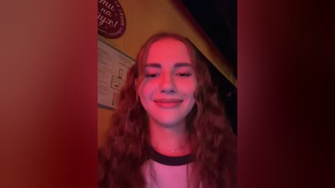 Video of a young woman with long, wavy brown hair, smiling broadly, standing indoors with a dimly lit, warm-colored background.