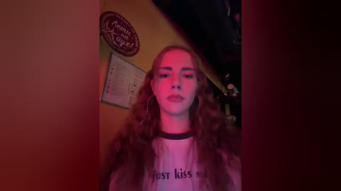 Video of a young woman with long curly brown hair, wearing a black choker, white T-shirt with \"fresh kiss\" written on it, standing indoors under dim red lighting.