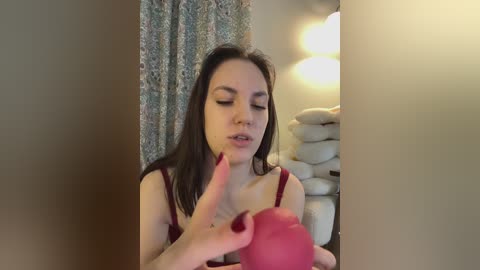 Video of a fair-skinned, dark-haired woman with a medium build and medium-sized breasts, wearing a red bra, holding a red apple in front of a beige wall and white pillows.