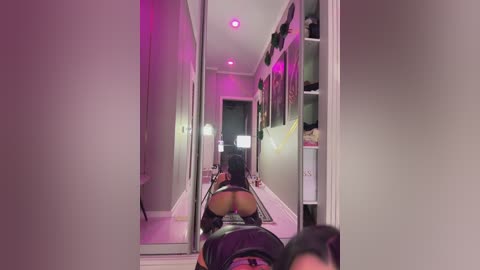 A video of a woman in black lingerie, kneeling on a floor mirror in a dimly lit hallway with pink lighting.