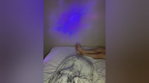 Video of a person lying on a bed, their feet pointing towards the camera, with a soft purple light casting a shadow on the wall. The bed has a white quilt with a faint grey pattern.