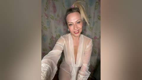 Video of a smiling blonde woman in a sheer white robe, revealing her breasts, standing against a floral-patterned curtain background.