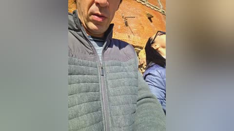 A video of a middle-aged man with a fair complexion, wearing a gray quilted jacket and a blue shirt, standing in front of a brick wall with a clock and a person partially visible in the background.