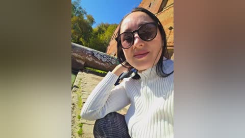 Video of a smiling, fair-skinned woman with straight black hair, wearing large sunglasses and a white ribbed sweater, standing on a cobblestone path beside a brick building with graffiti, under a bright blue sky.