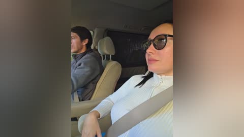 A video of a woman with black sunglasses and a white sweater, driving a car, while a man with a beard sits in the passenger seat.