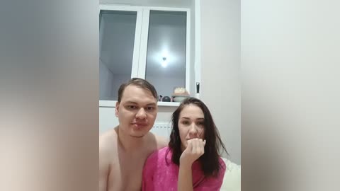 Video of a young Caucasian couple, both shirtless, smiling. The woman, with long dark hair, is wearing a pink robe. They are in a white-walled room with a window and a shelf in the background.
