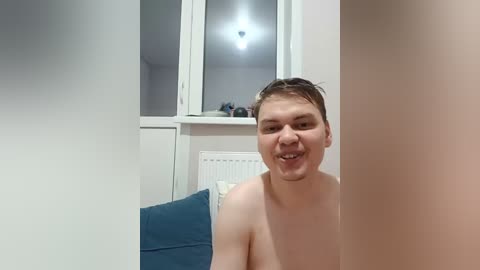 A video of a shirtless, fair-skinned young man with short brown hair, smiling, standing in a modern, minimalist room with white walls, a radiator, and a blue cushion.